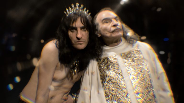 Two performers in glittering costumes on stage, one wearing a spiked crown, lit with chromatic stage lighting.