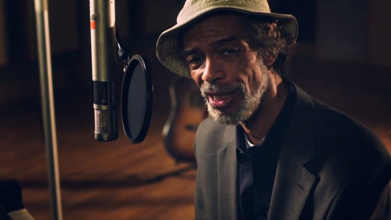 Gil Scott-Heron speaking into a studio microphone, guitar blurred behind him.