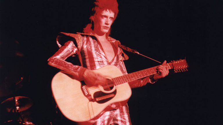 A glam performer in a sequined jumpsuit with a red mullet, playing a 12-string acoustic guitar under red stage lighting.
