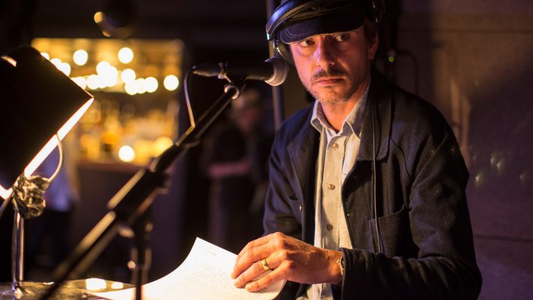 A man in a flat cap and headphones reading into a microphone at a lit desk, bar lights blurred behind him.