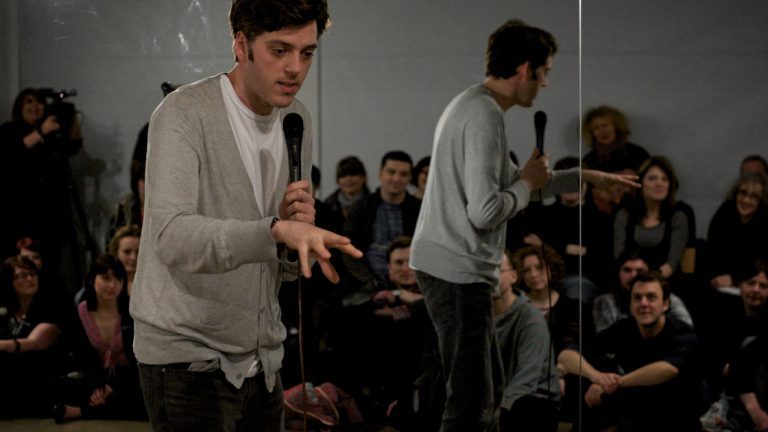 A man in a grey cardigan performing into a microphone before a seated audience, in a double-exposed image.