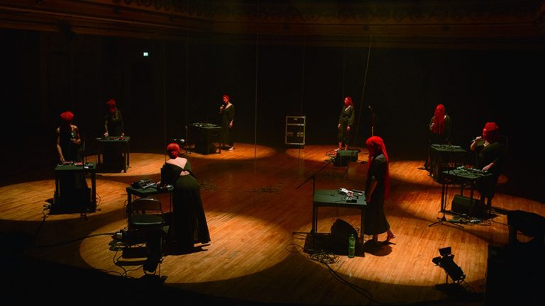 Gazelle Twin & NYX performers in red hoods and dark robes at electronics stations.